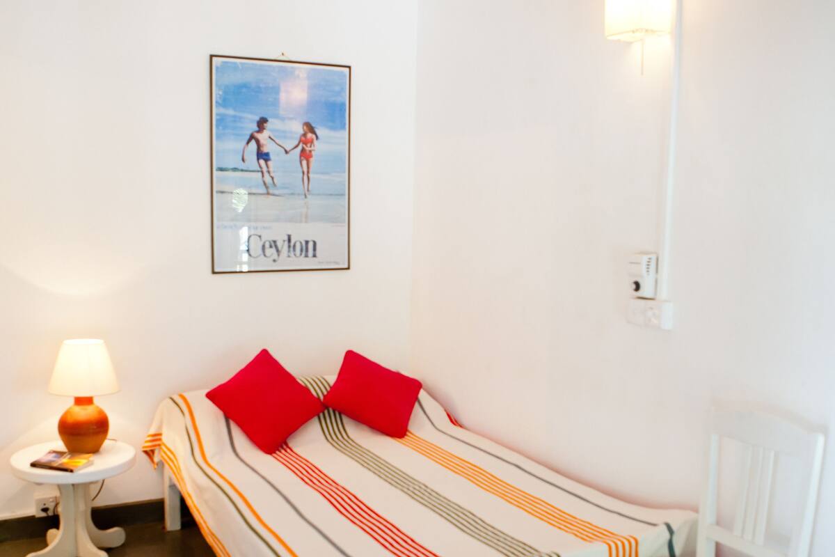 A neatly arranged bedroom with a single bed, a chair, a small table, a lamp, a framed picture on the wall, and a ceiling fan.