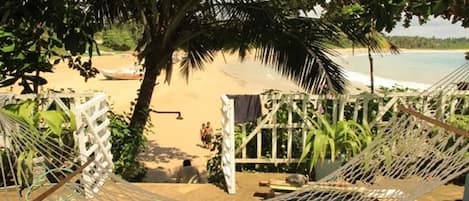 Una hamaca atada entre dos postes blancos, con vista a una playa arenosa donde se ven algunas personas a lo lejos y un barco en el agua.