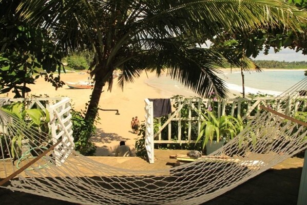 A hammock tied between two white posts, overlooking a sandy beach with a few people in the distance and a boat on the water.