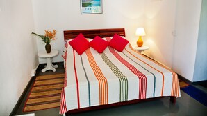 A neatly made bed with a striped bedspread, a small bedside table with a lamp, and a wall-mounted poster. - Talalla Bay Beach House (Talalla)