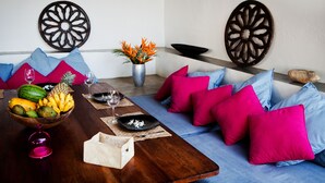 A dining area with a wooden table, colorful cushions, decorative wall art, and a fruit bowl. - Talalla Bay Beach House (Talalla)