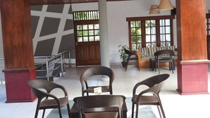 A spacious lobby with a high ceiling, wooden columns, and a staircase. There are wicker chairs and a sofa with a view of greenery outside. - Paradise Inn (Kandy)