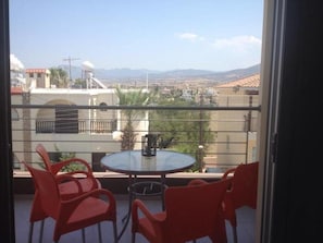 A balcony with a round table and red chairs, overlooking a cityscape with buildings and mountains.