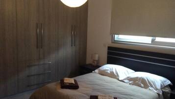 A bedroom with a bed, a nightstand, a lamp, a wardrobe, and a window with blinds.