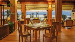 Uma área de jantar de madeira com uma mesa posta para quatro pessoas, rodeada por cadeiras, e vista para um corpo d'água com montanhas ao fundo.