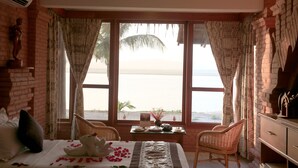 A hotel room with a bed, two wicker chairs, a small table with a vase, and a view of the beach through large windows. - KMA Bagan Hotel River View (Nyaung-U)