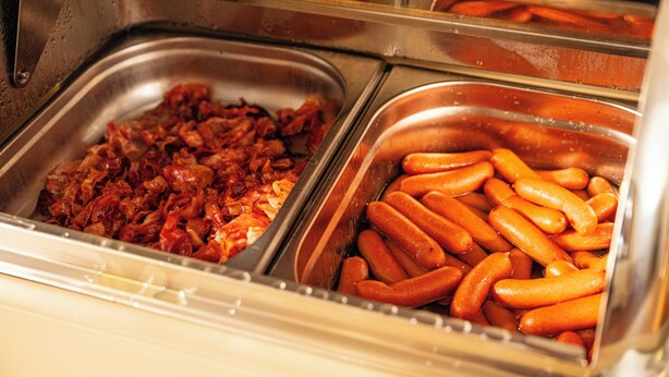 A buffet with two trays of food, one with what appears to be bacon and the other with sausages. - Hotel Adalbert (Prague)