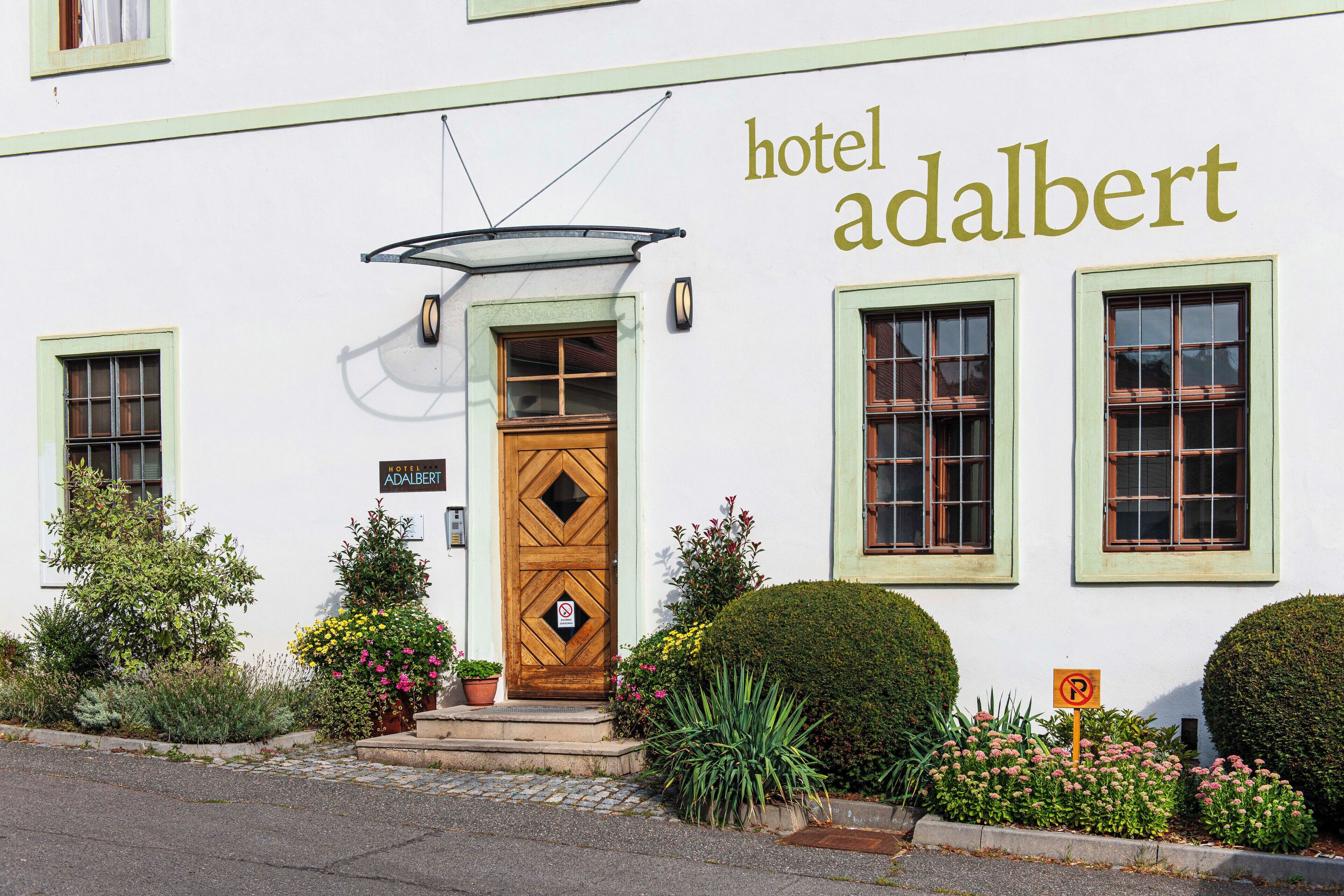 A hotel named 'hotel adalbert' with a wooden door, a sign indicating no parking, and a small garden with various plants and flowers.
