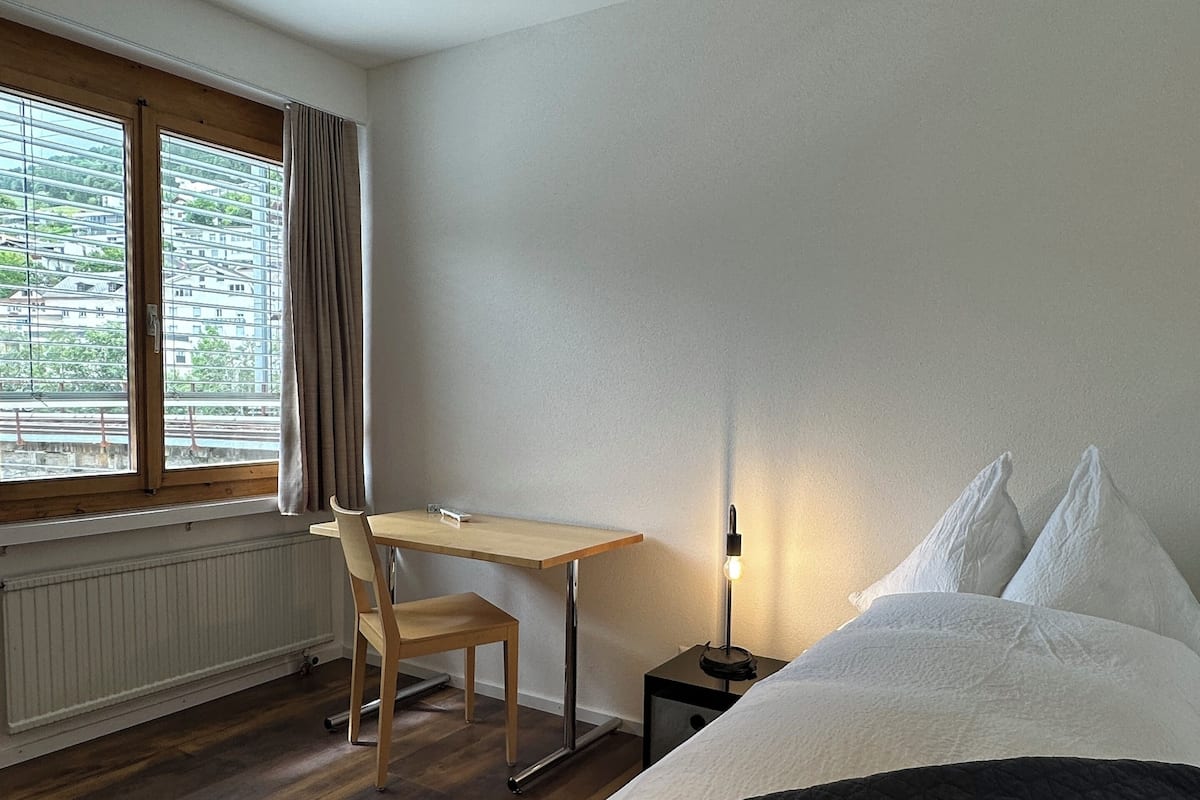 A hotel room with a bed, a desk, and a chair. There is a window with curtains and a view of buildings outside.