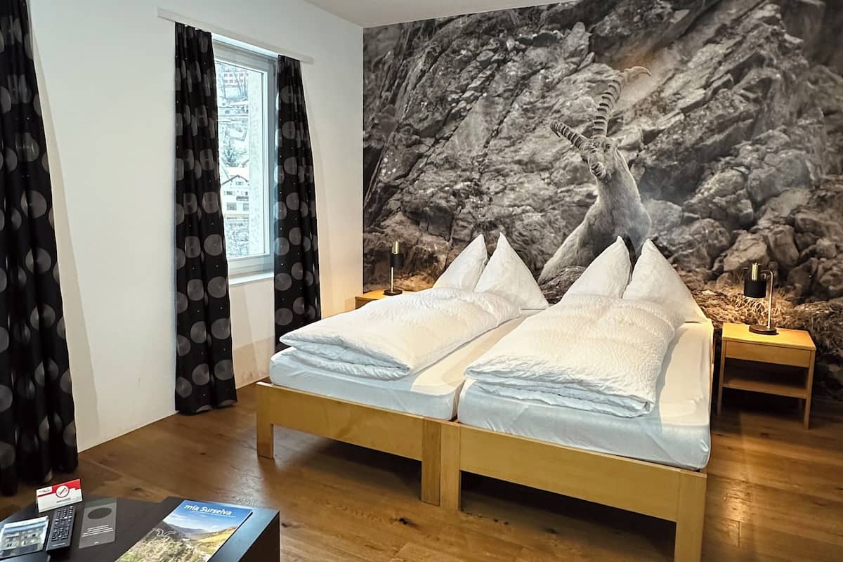 A double bed with white linens, a wooden headboard, a small bedside table with a lamp, a large wall mural of a rocky landscape, and a window with black curtains.