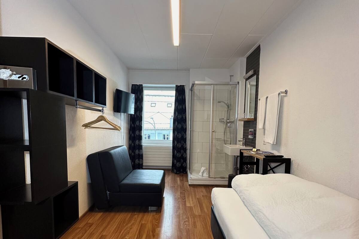 A modern hotel room with a wooden floor, a black sofa, a white bed, a glass-enclosed shower, a TV, and a wardrobe.