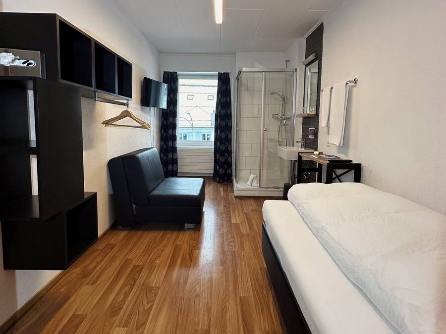 A modern hotel room with a wooden floor, a black sofa, a white bed, a glass-enclosed shower, a TV, and a wardrobe.