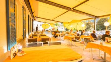 Un café en plein air avec des tables et des parasols jaunes, et une rangée de fenêtres dotées de volets.