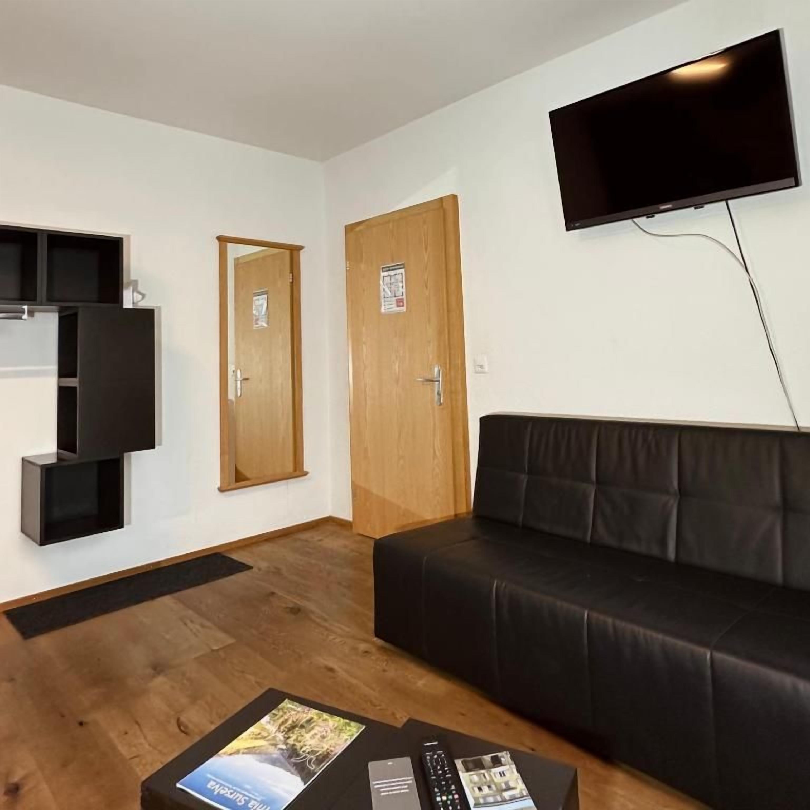 Une pièce avec un canapé en cuir noir, un téléviseur à écran plat fixé au mur, une porte en bois et une petite table sur laquelle se trouve une télécommande.