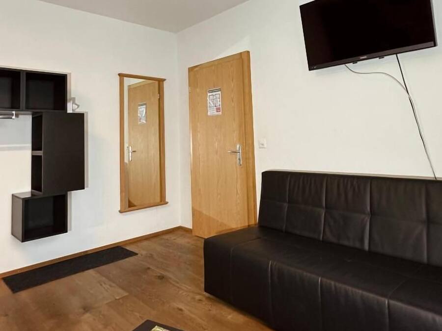 Una habitación con un sofá de cuero negro, un televisor de pantalla plana montado en la pared, una puerta de madera y una pequeña mesa con un control remoto.