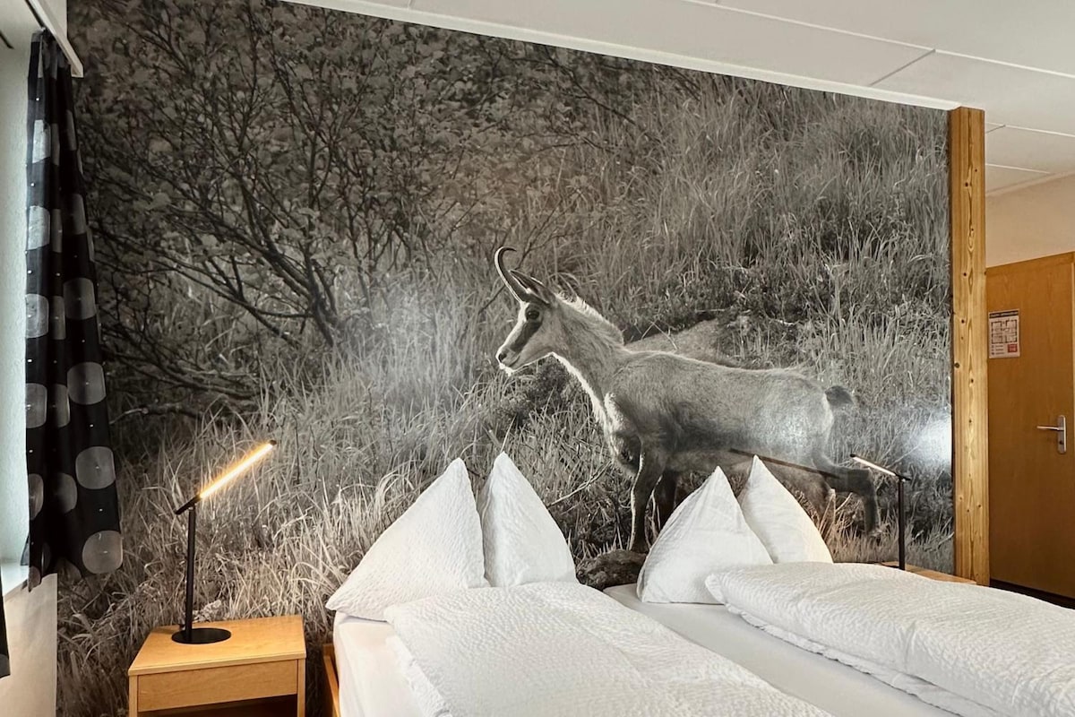 Un dormitorio con dos camas, una mesita de noche, una lámpara y un gran mural en la pared con un ciervo.