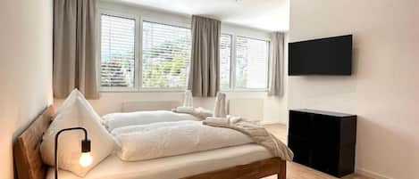 A modern bedroom with a wooden bed, a bedside lamp, a flat-screen TV, and a window with curtains.