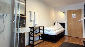 Habitación de hotel compacta con ducha, cama, una mesita y suelo de madera.
