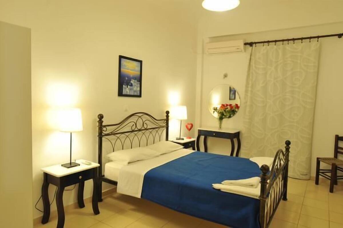 Un dormitorio ordenado con una cama, dos mesitas de noche con lámparas, una silla y una ventana con cortina.