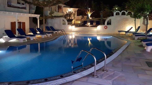 A nighttime swimming pool with lounge chairs and a lifebuoy, surrounded by white buildings with balconies and palm trees. - Sunflower Santorini (Santorini)