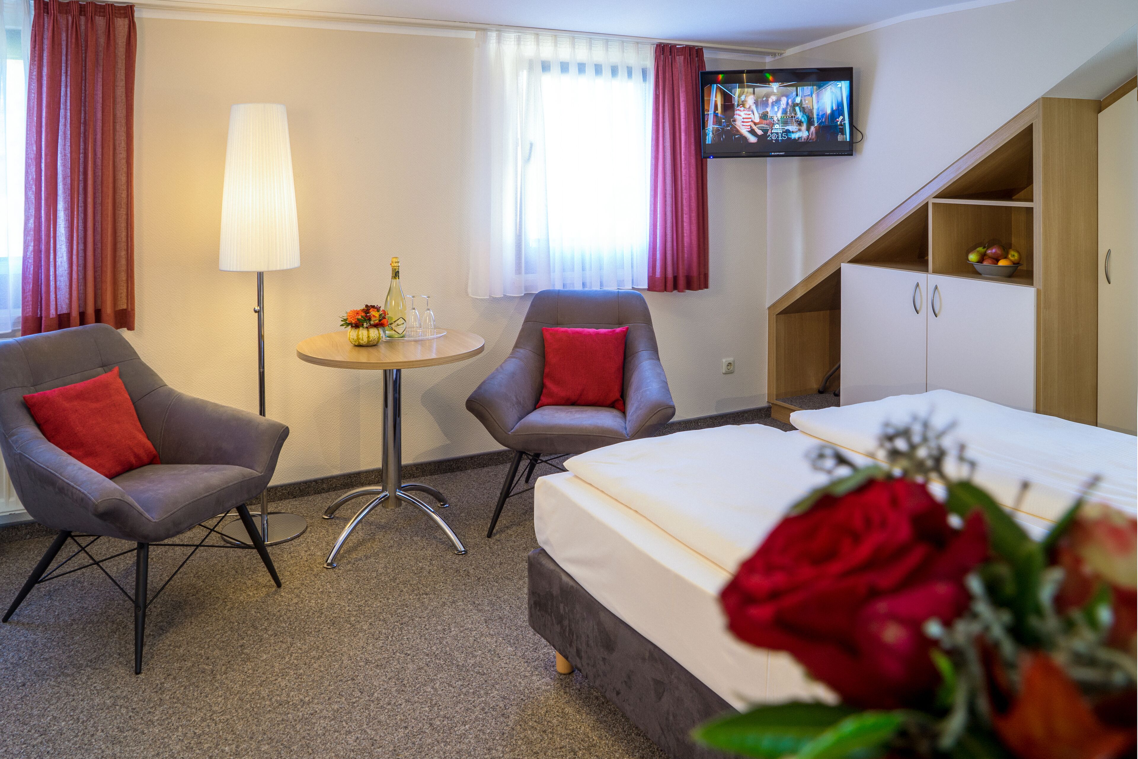 A hotel room with a bed, two armchairs, a small table with a lamp, a TV on the wall, and a staircase leading to a loft area.