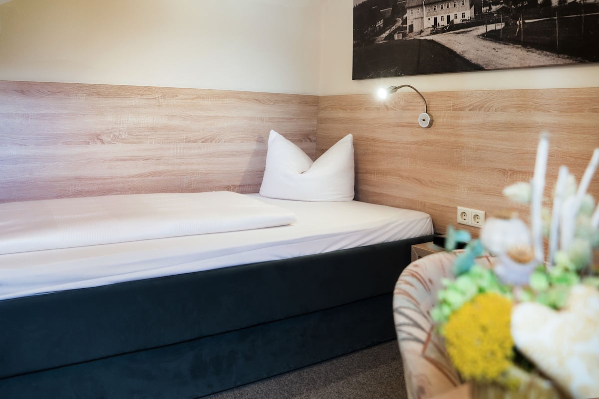 A modern hotel room with a wooden headboard, a single bed with white bedding, a bedside table, and a wall-mounted lamp.