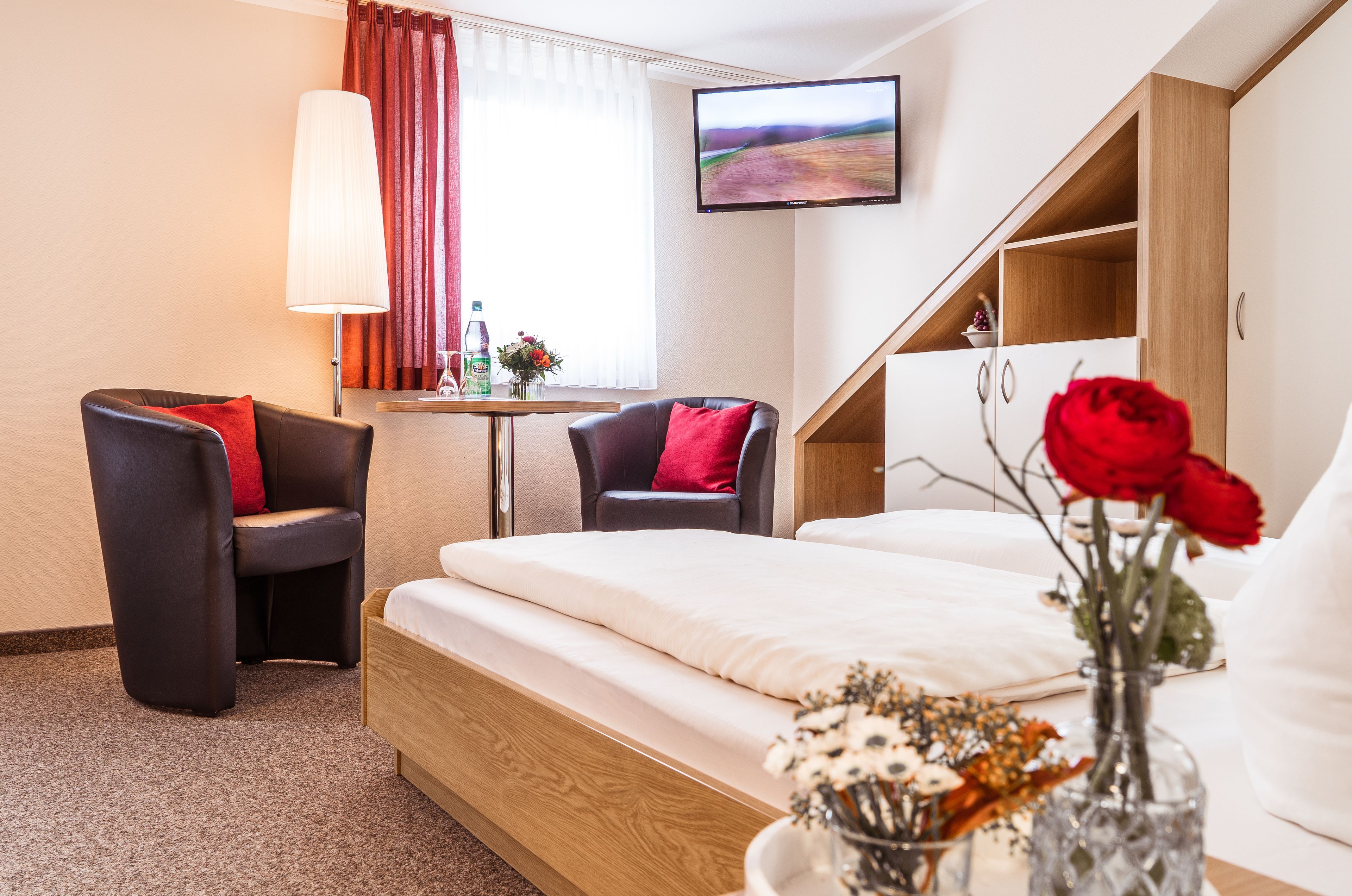 A hotel room with a bed, two armchairs, a small table with a lamp, a TV on the wall, and a vase with flowers.