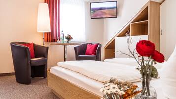 A hotel room with a bed, two armchairs, a small table with a lamp, a TV on the wall, and a vase with flowers.