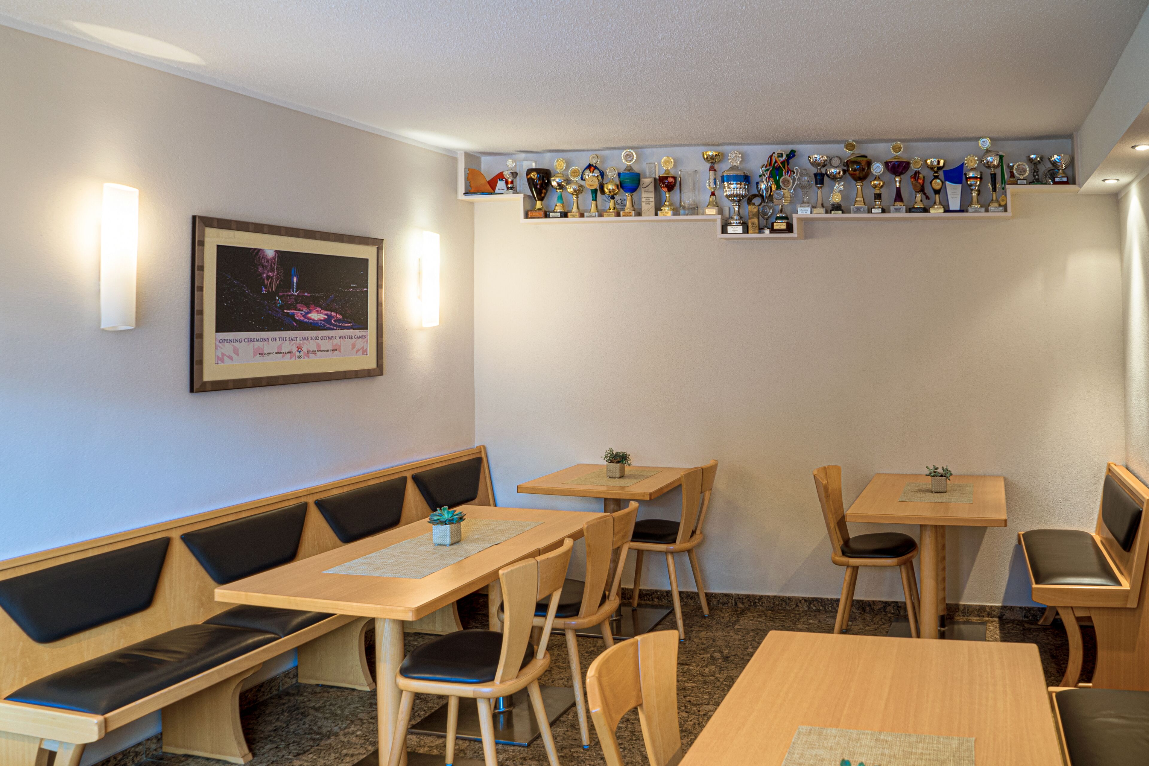 Ein Restaurant mit Holztischen und -bänken, einem Wandregal mit Pokalen und einem gerahmten Bild an der Wand.