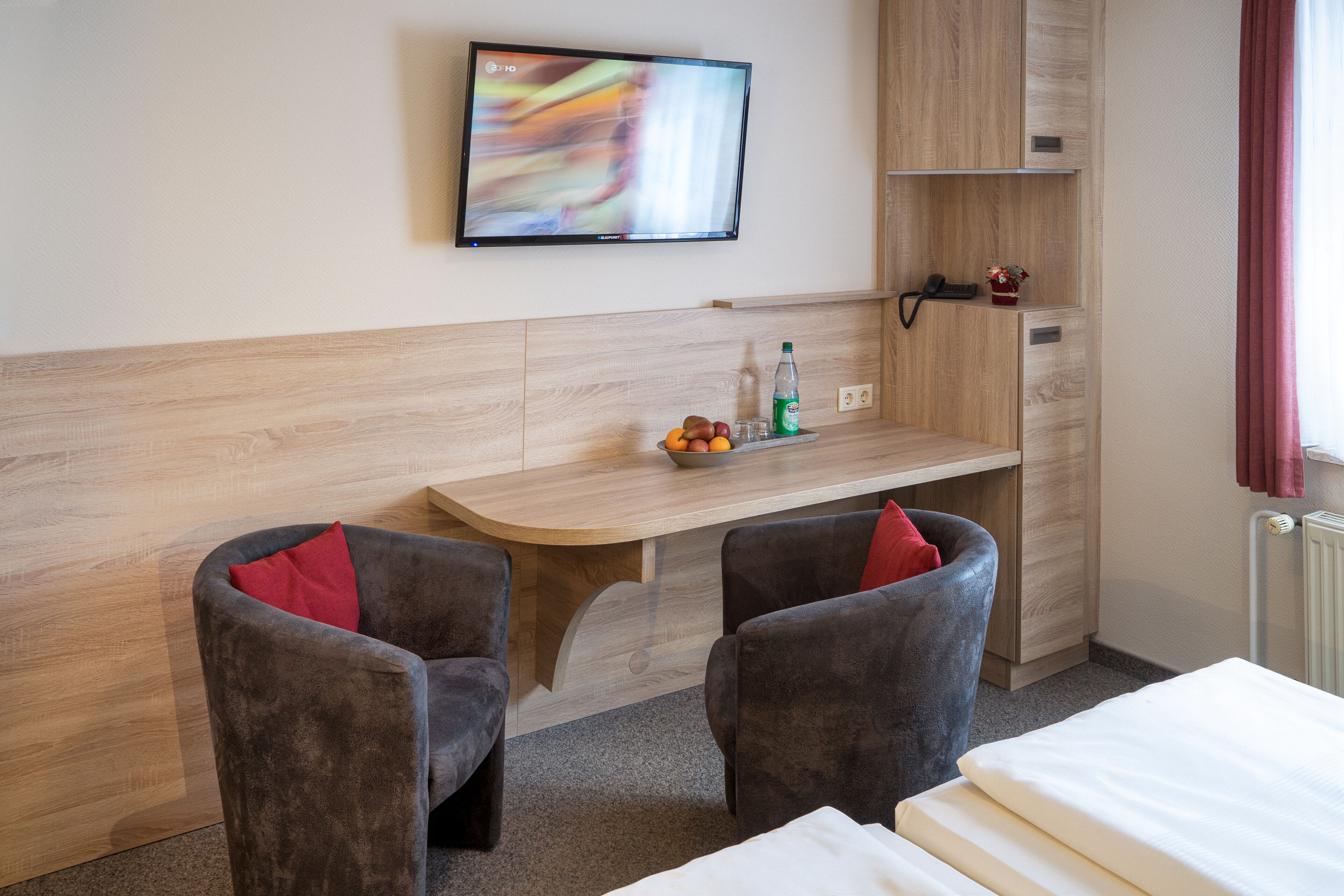 Ein Hotelzimmer mit an der Wand befestigtem Fernseher, einem Schreibtisch mit Obstschale, zwei Sesseln und einem Bett.