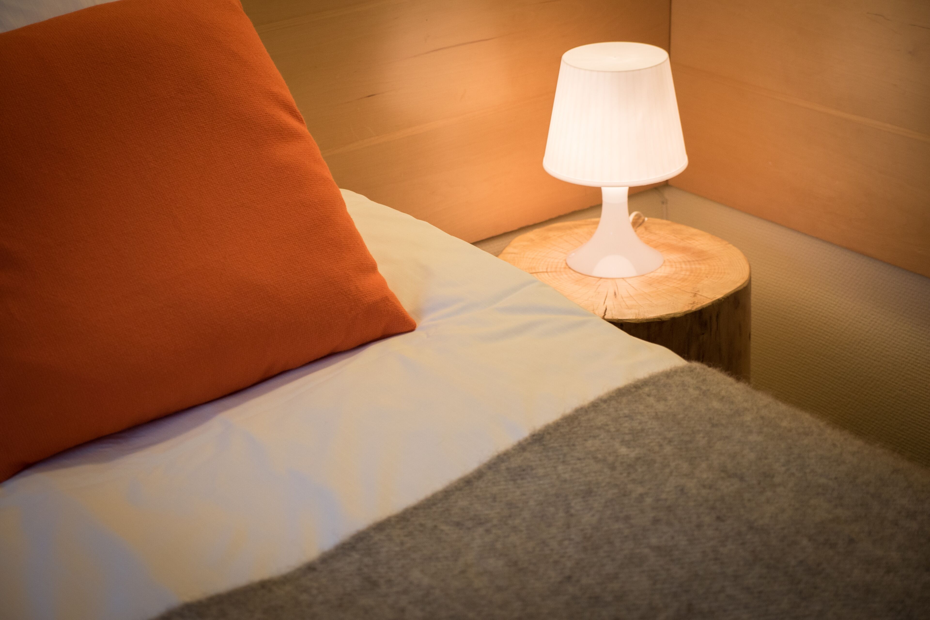 En snyggt bäddad säng med en vit och orange kudde, en grå pläd och en vit bordslampa på ett träbord bredvid sängen.