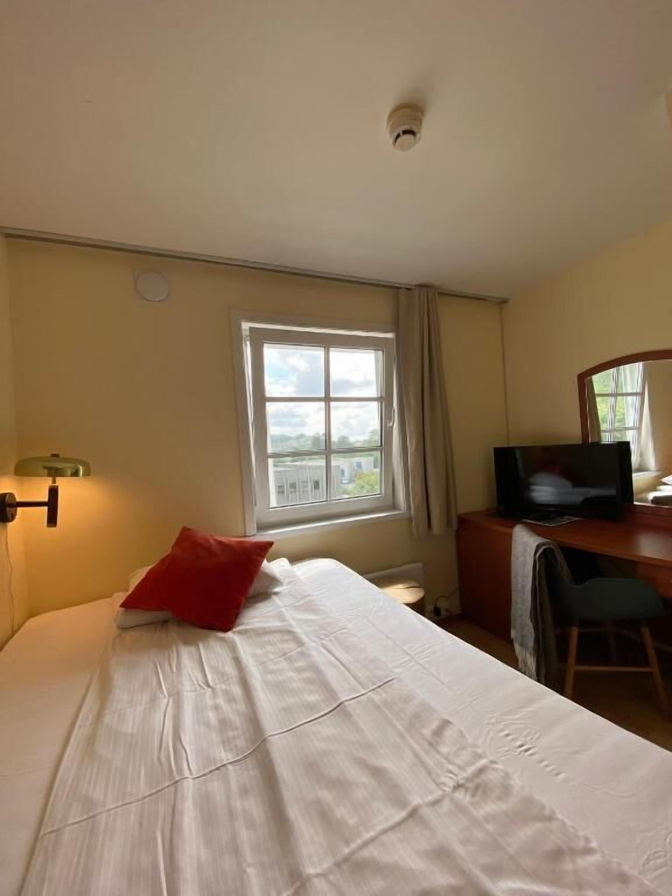 A hotel room with a bed, a desk with a television, a window with curtains, and a ceiling light.