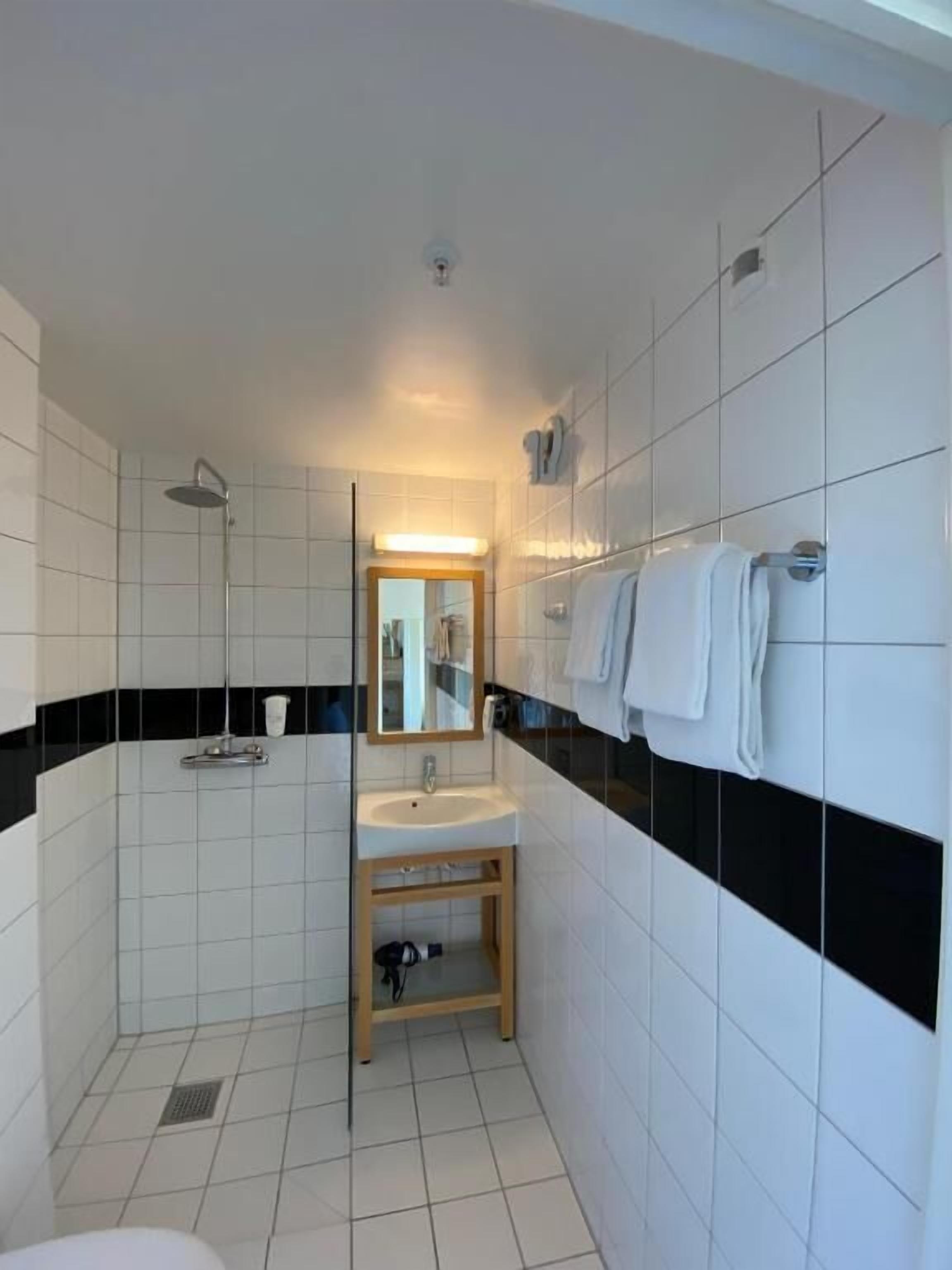 A small, clean bathroom with white tiles, a sink, a mirror, and a shower.