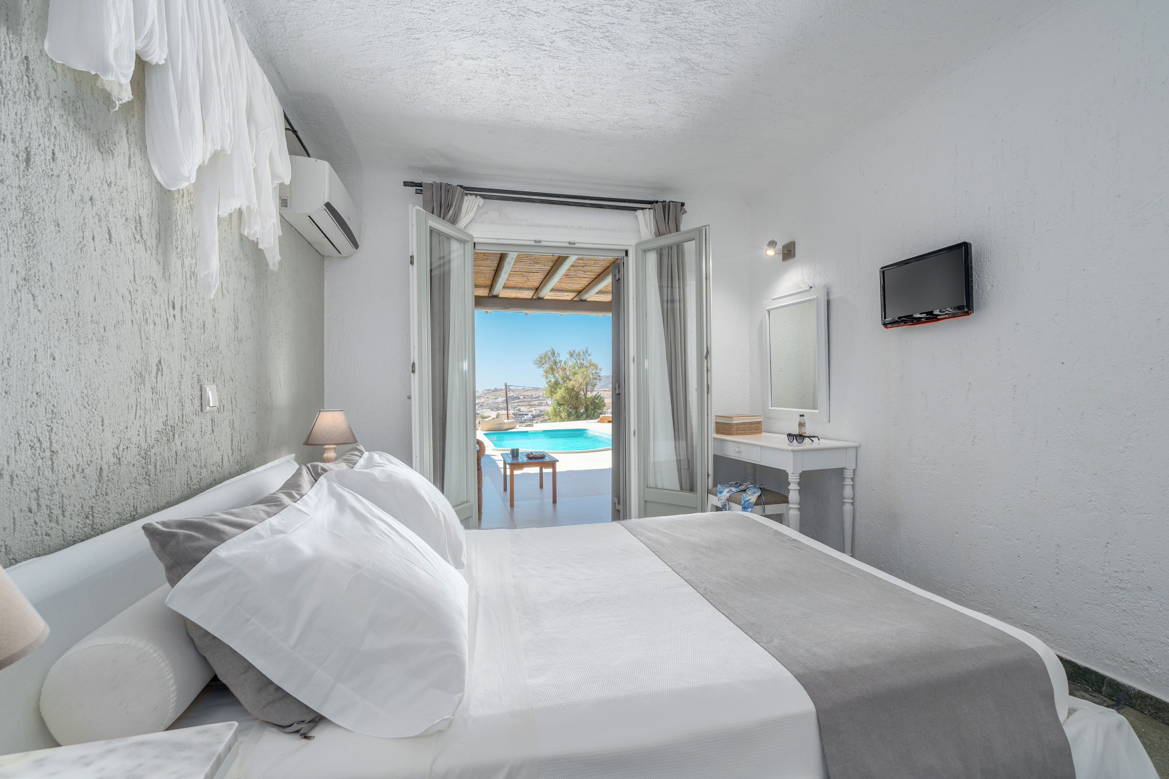 A hotel room with a bed, a bedside table with a lamp, a bathroom with a mirror, and a view of a pool.