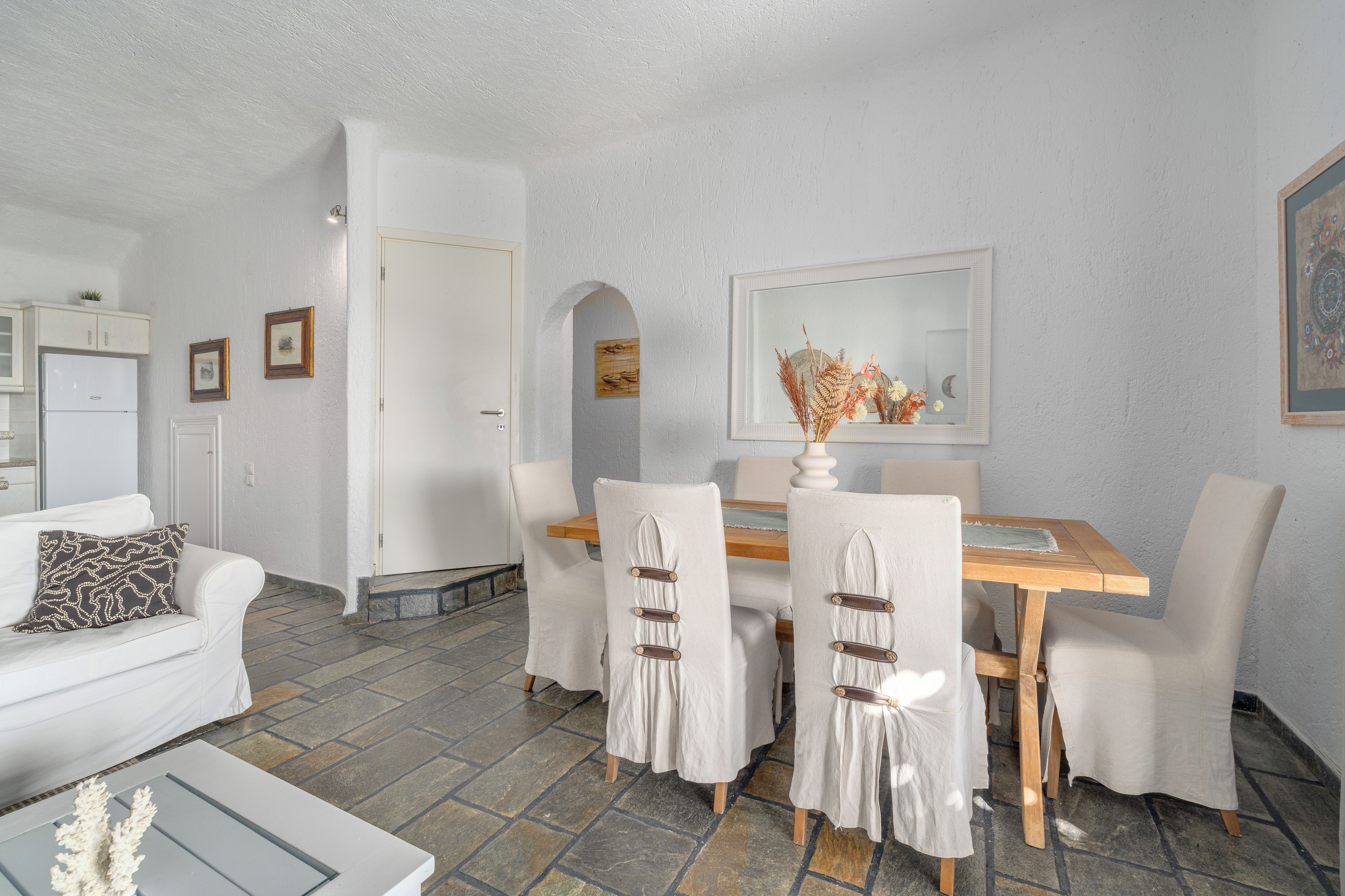 Un coin repas avec une table en bois et des chaises blanches, un canapé blanc agrémenté d’un coussin à motifs, et une cuisine en arrière-plan.
