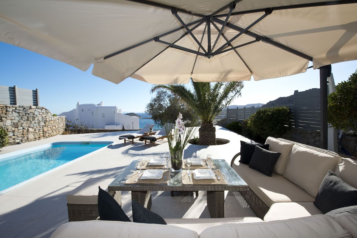 A poolside area with a table set for four, a white umbrella, and a view of the sea and buildings in the background.