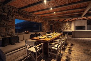 A dining area with a stone wall, wooden ceiling, and a large window overlooking a mountainous landscape. - Thalasses Villas (Mykonos)