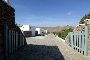 A stone-paved street leading to white buildings with a stone wall and a metal gate. - Thalasses Villas (Mykonos)