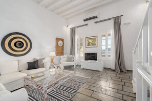 A living room with a white sofa, a glass coffee table, a mounted clock, a painting, and a TV on a white stand. - Thalasses Villas (Mykonos)