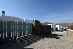 A stone-paved courtyard with a green gate, white buildings, and a clear blue sky. - Thalasses Villas (Mykonos)