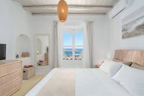A bedroom with a bed, a TV, a dresser, a mirror, and a view of the sea. - Thalasses Villas (Mykonos)