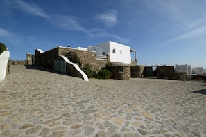 A stone-paved courtyard with a white building featuring a stone facade and a white fence. - Thalasses Villas (Mykonos)