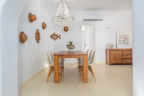 A dining area with a wooden table, white chairs, a chandelier, wall art, and a white door. - Thalasses Villas (Mykonos)