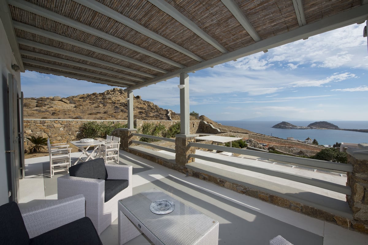 A covered outdoor seating area with white wicker furniture, overlooking a coastal landscape with rocky hills and the sea.