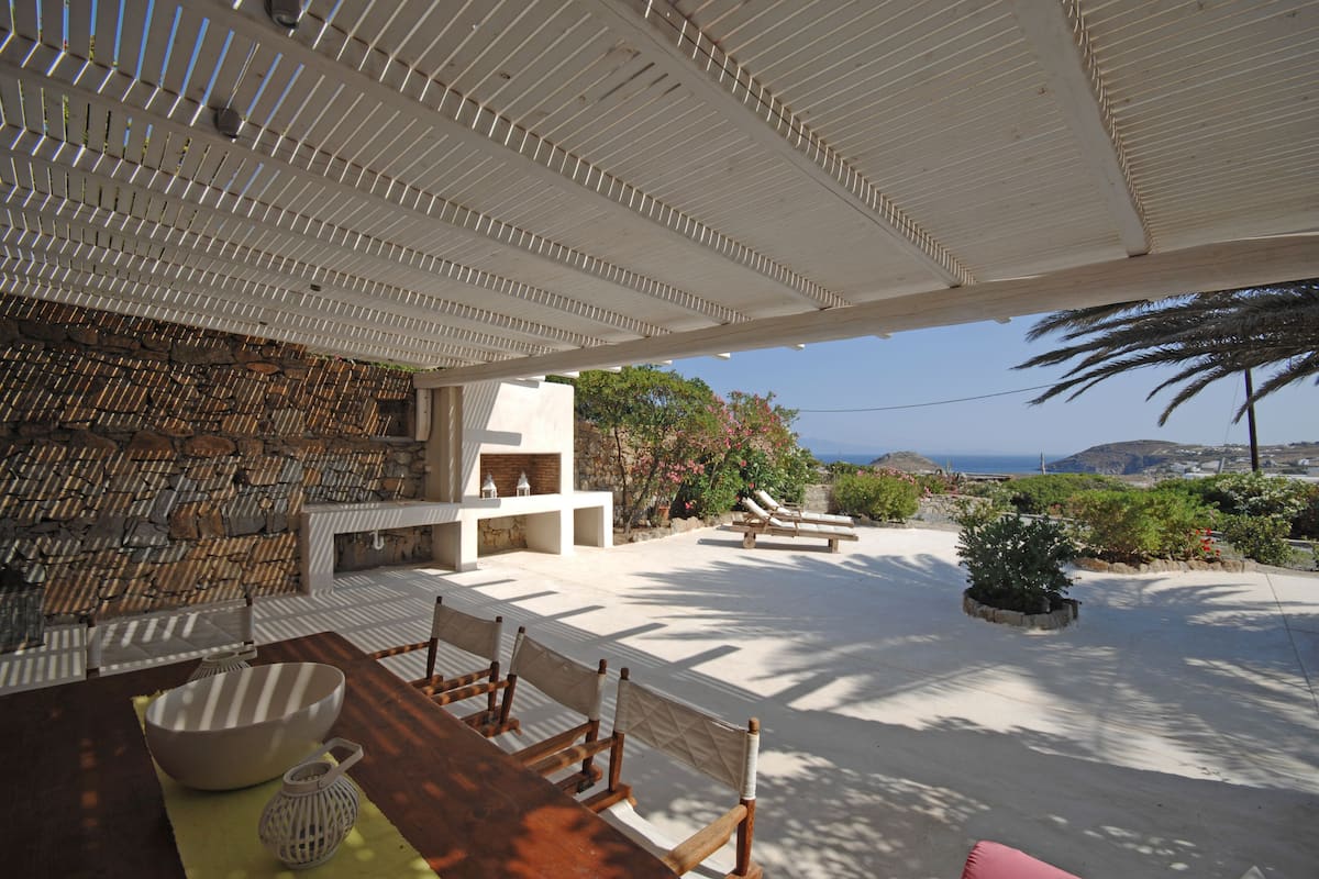An outdoor patio with a stone wall, a table with chairs, a white grill, and a view of the sea.