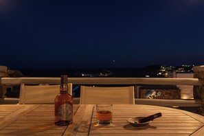 A bottle of Chivas Regal whisky, a glass of whisky, and a cigar on a wooden table with a view of the city at night. - Thalasses Villas (Mykonos)