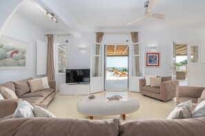 A spacious living room with a flat-screen TV, beige sofas, a white coffee table, and a ceiling fan. - Thalasses Villas (Mykonos)