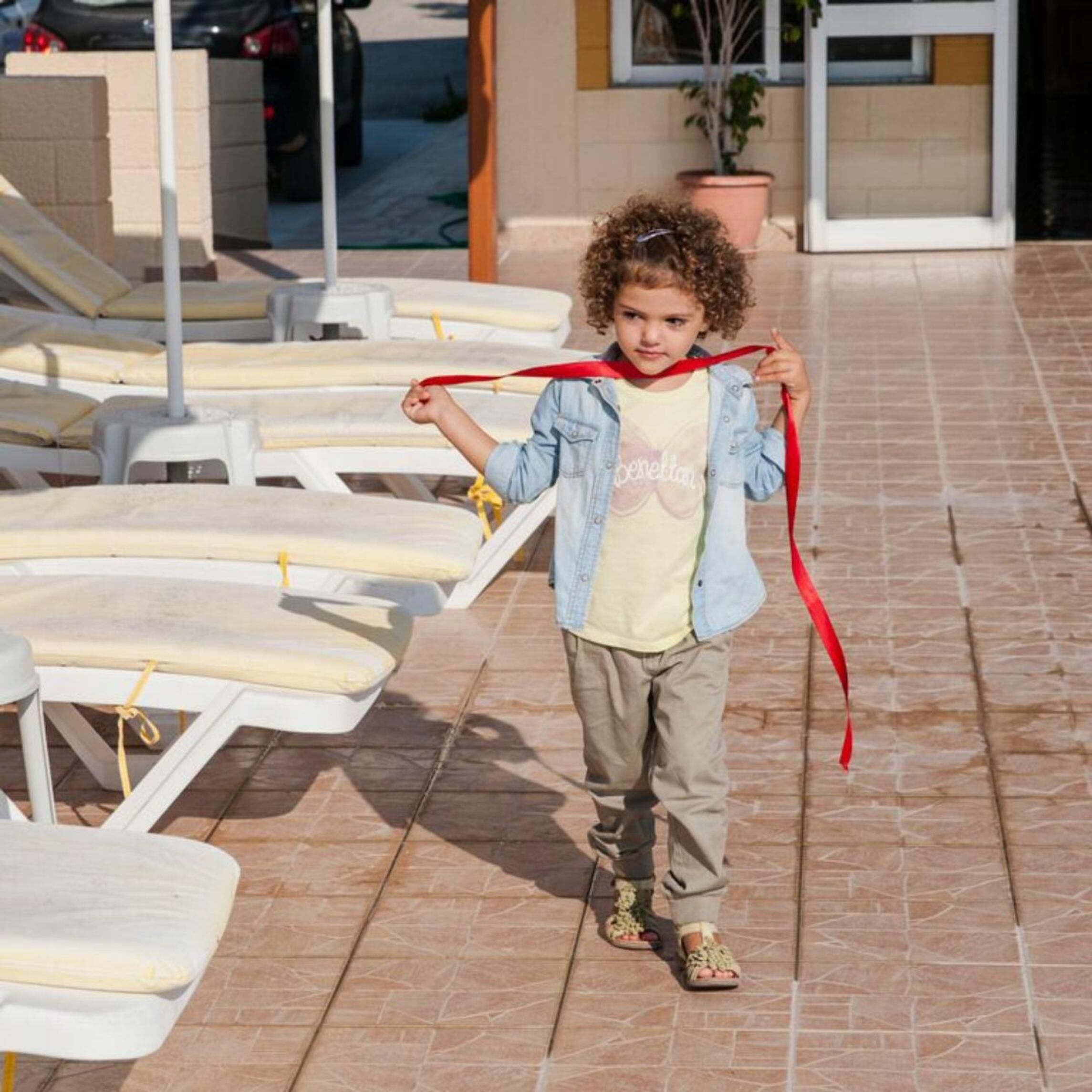 Un enfant tenant un ruban rouge, debout sur une terrasse carrelée avec des chaises longues.
