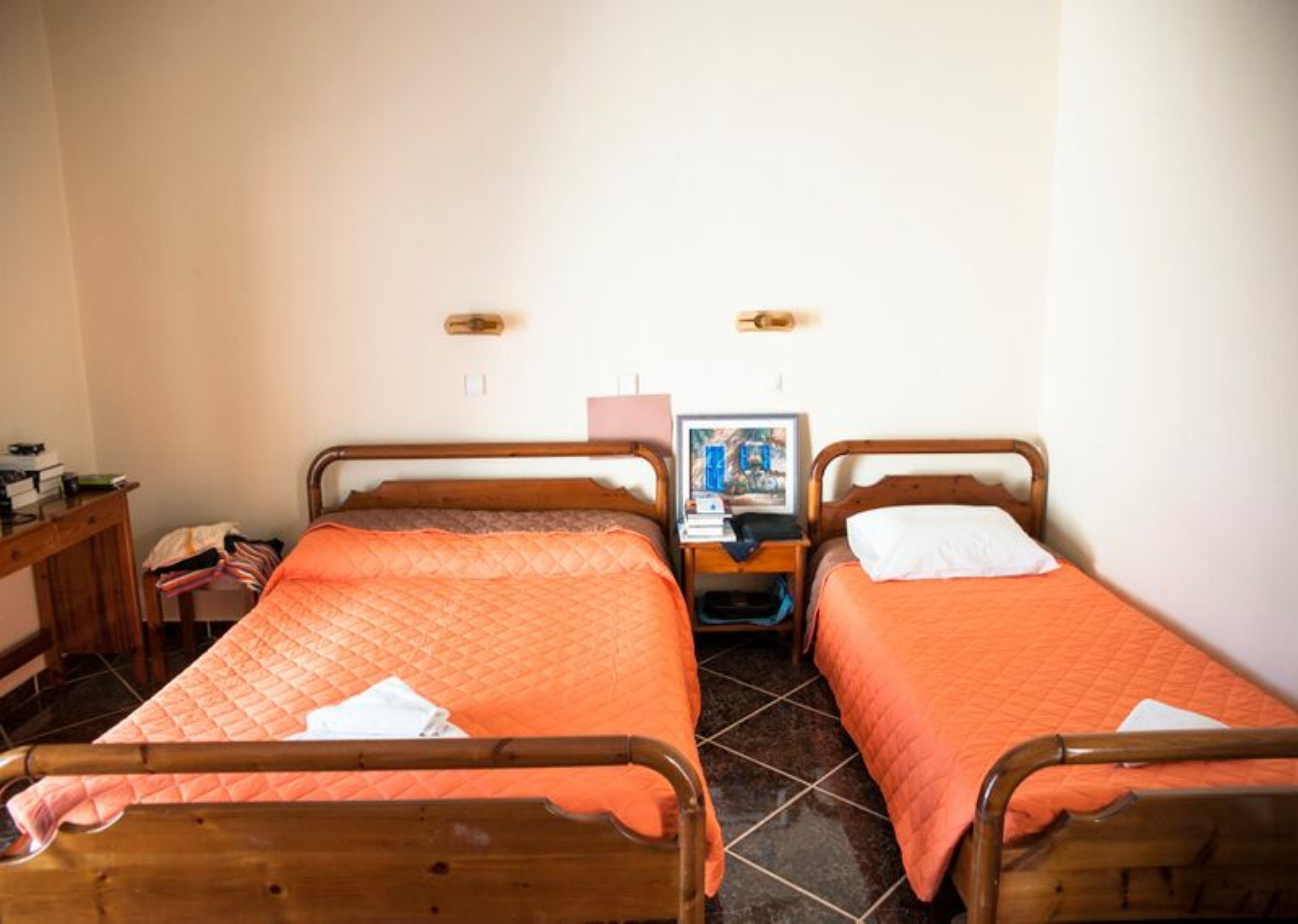 A room with two single beds, each with an orange quilted bedspread and white pillows. There is a wooden nightstand with a lamp and a framed picture between the beds. The room has tiled flooring and a wooden desk with a chair to the left.