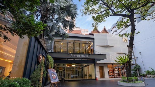 The exterior of the Ariston Hotel with a modern facade, large glass entrance, and surrounding greenery. - Ariston Hotel (Bangkok)